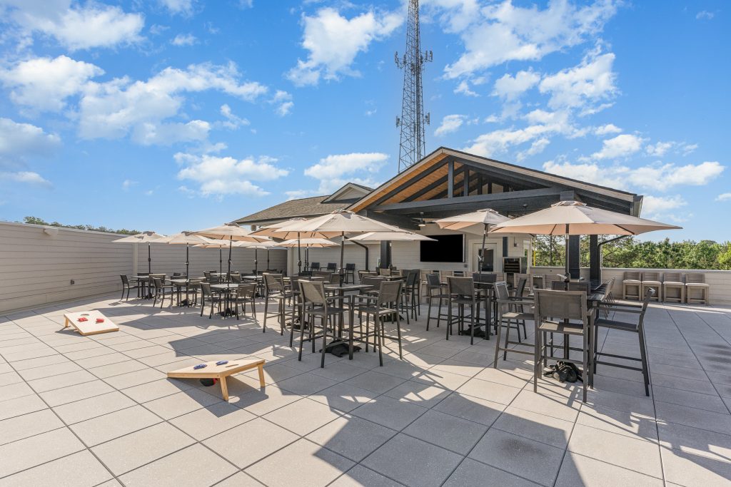 Experience the spacious rooftop patio featuring comfortable seating, shaded tables with umbrellas, and recreational activities like cornhole. Perfect for outdoor gatherings and relaxation.